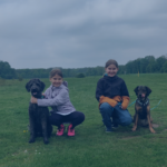 Zwei Mädchen hocken entspannt auf einer grünen Wiese neben einem schwarzen lockigen Hund und einem braun-schwarzen Mischlingshund unter bewölktem Himmel.